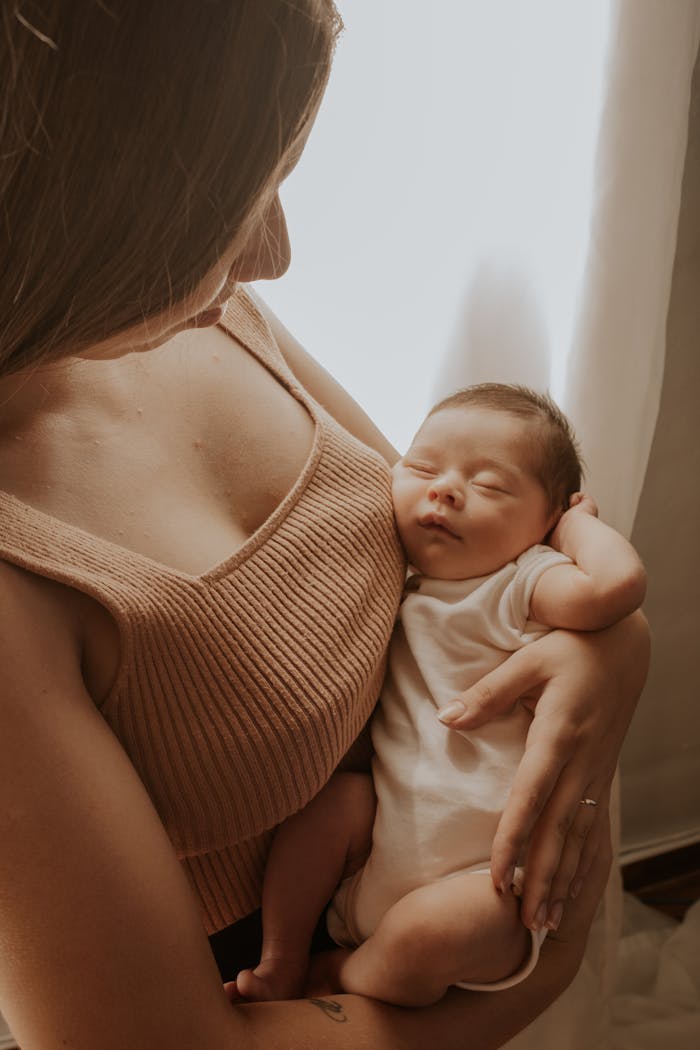 Mother lovingly holding her sleeping newborn child by a window with soft lighting.