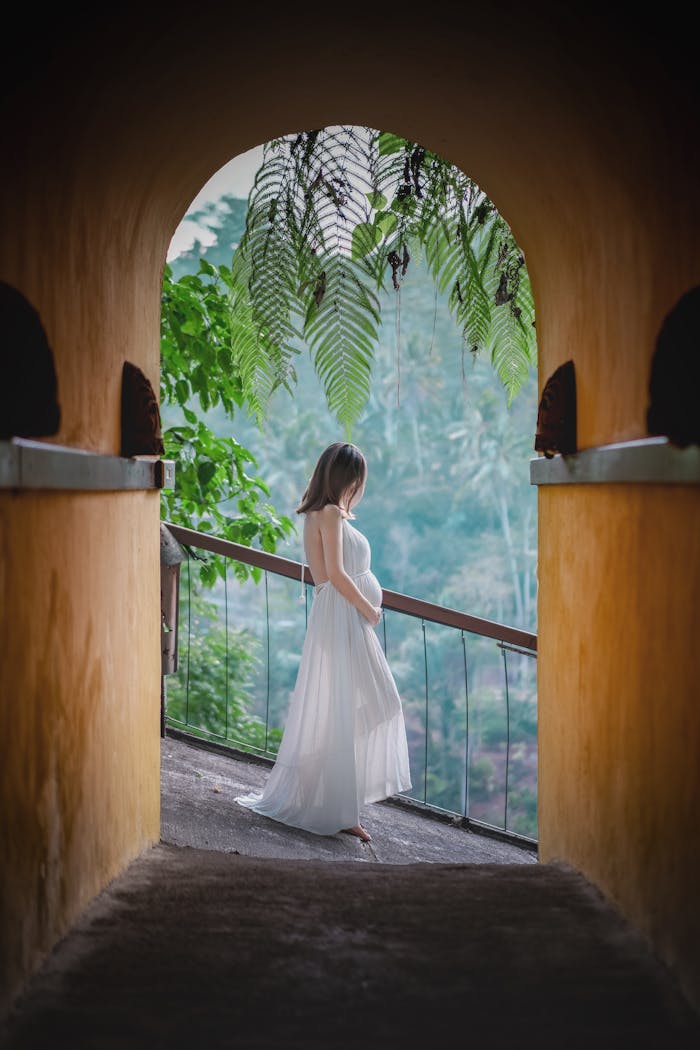 Pregnant woman in white dress stands peacefully in a lush tropical archway.